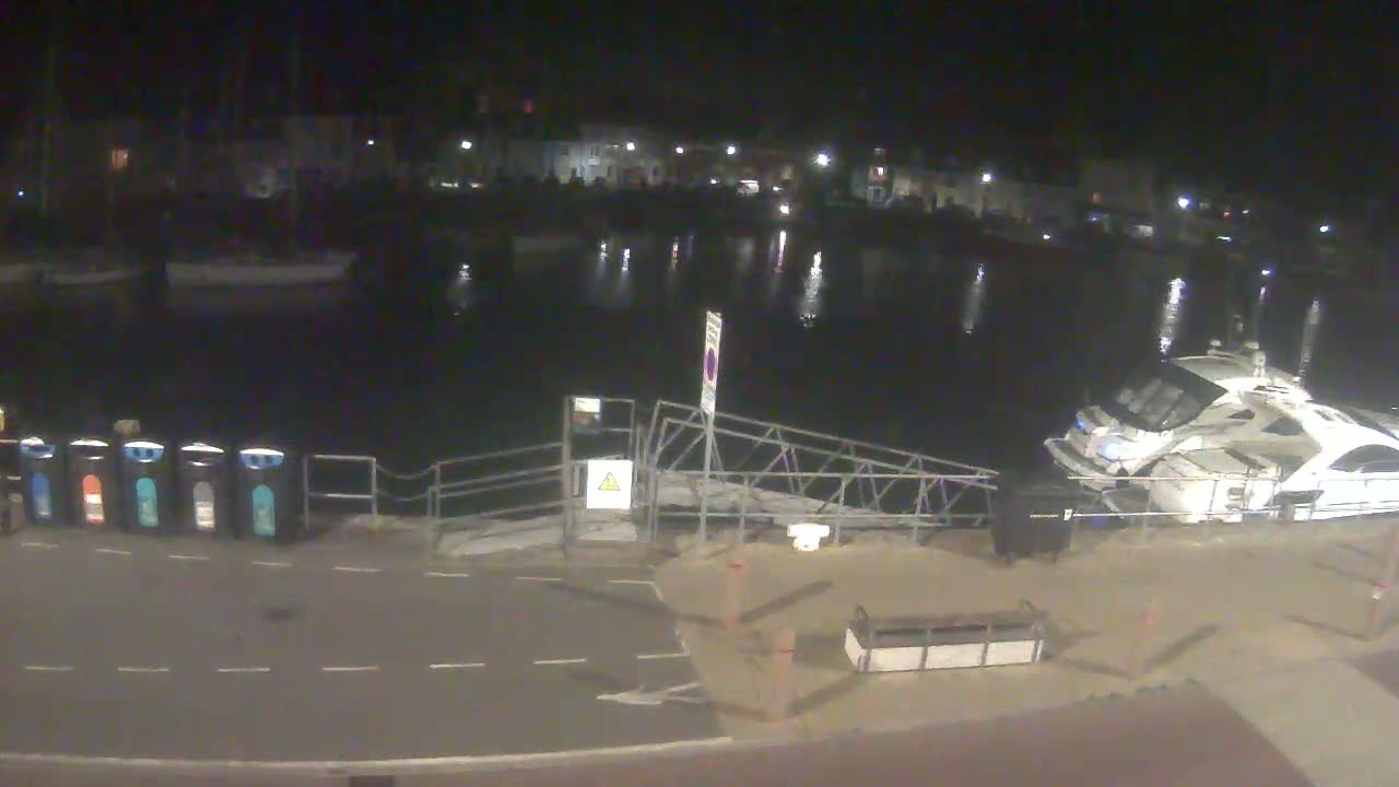 A nighttime view of a calm harbor with several boats, a small dock, and a row of trash cans.