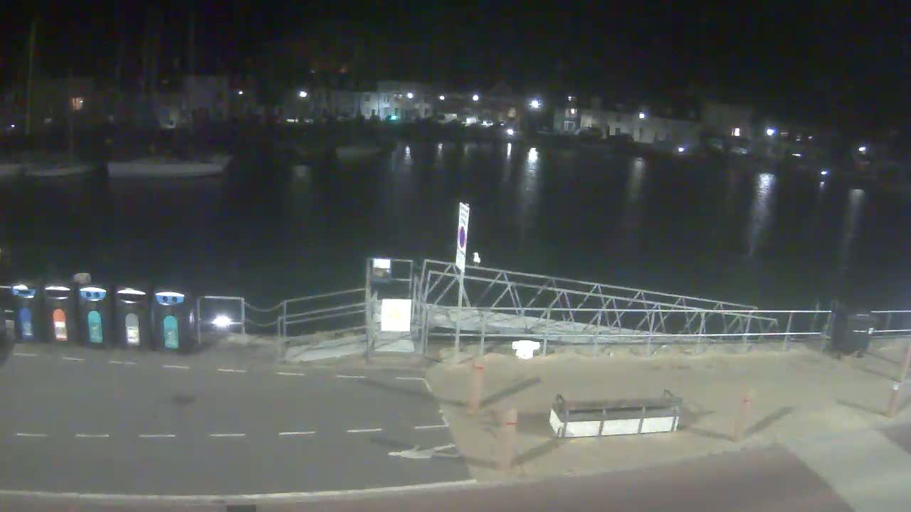 A nighttime view of a calm harbor with boats moored, a metal gangway leading to the water, and several recycling bins along a paved area.