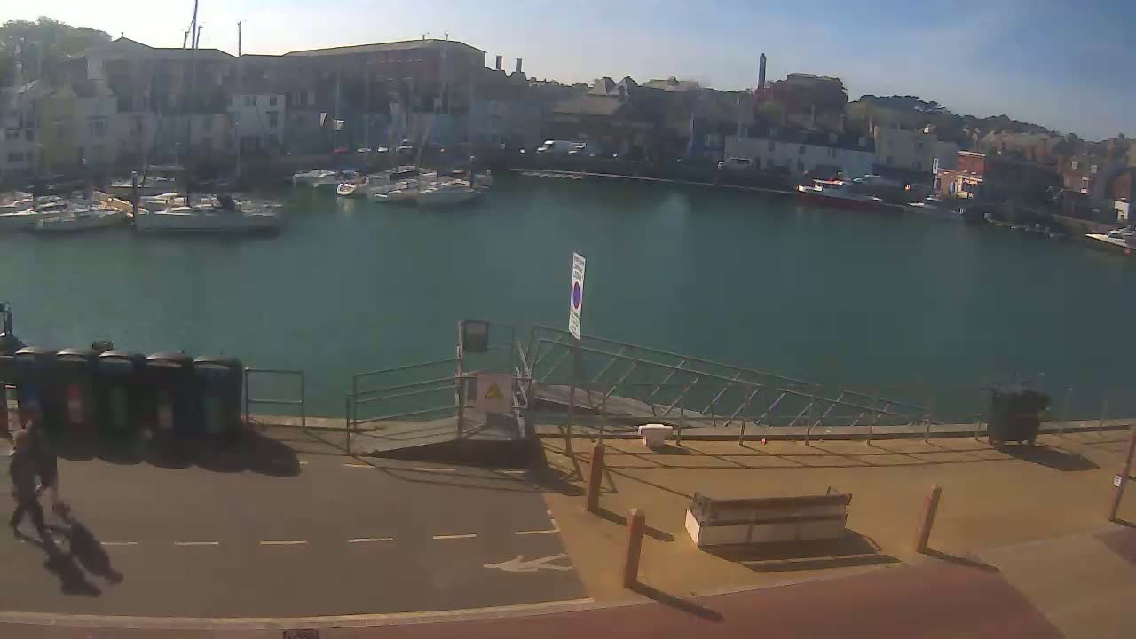 A calm, sunny day reveals a harbor filled with sailboats and buildings along the waterfront, with two people walking near a paved area.