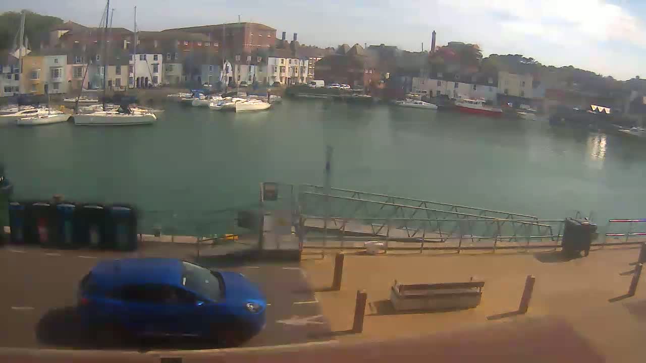 A blue car is parked near a harbor filled with sailboats on a partly sunny day.