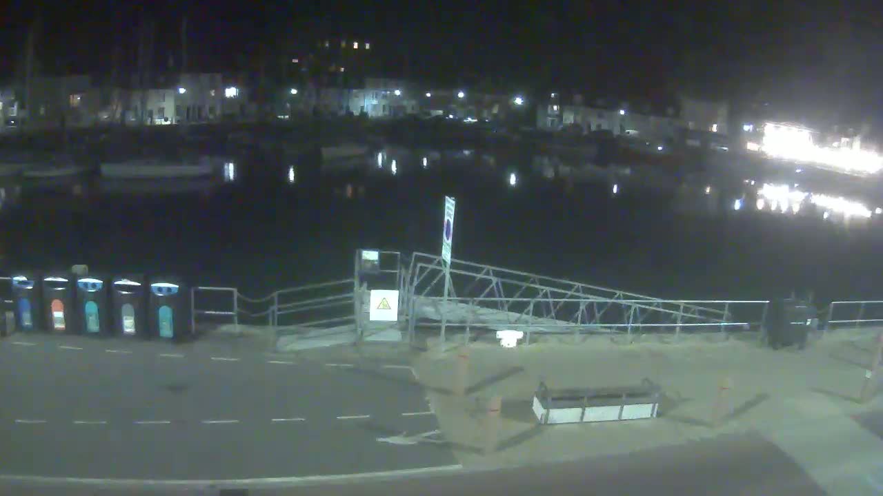 At night, a calm harbor is seen with boats moored along a waterfront, a metal gangway leading to the shore next to a row of recycling bins.
