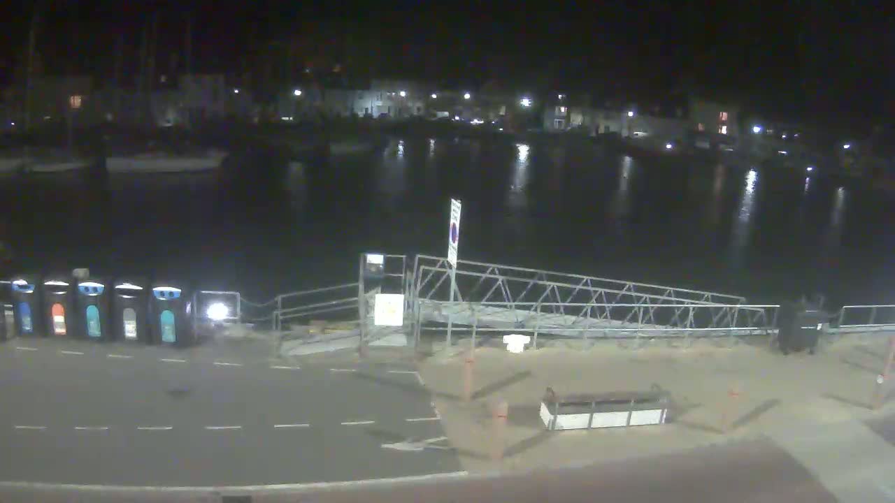 At night, a calm body of water reflects the lights of buildings across a small harbor, next to a metal gangway leading to a paved area with several recycling bins.
