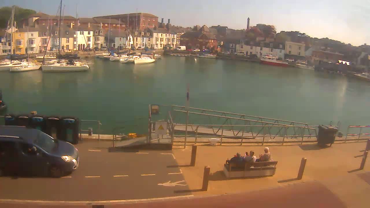 A harbor filled with sailboats is overlooked by colorful buildings under a sunny sky; a small group of people sit on a bench near a ramp leading to the water.