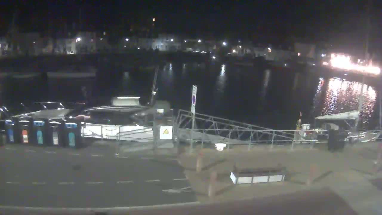 A nighttime view of a harbor shows several boats docked along a pier, under calm, dark water.
