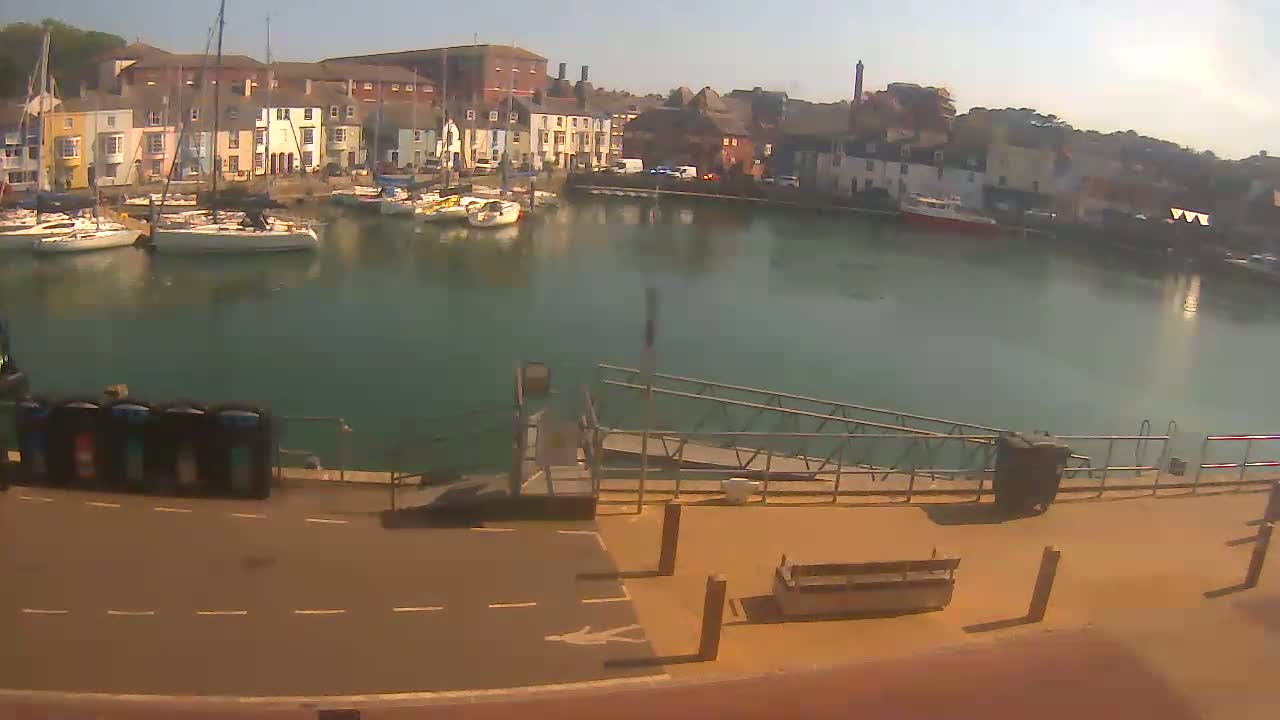 A calm harbor filled with sailboats sits before brightly colored buildings under a sunny sky.