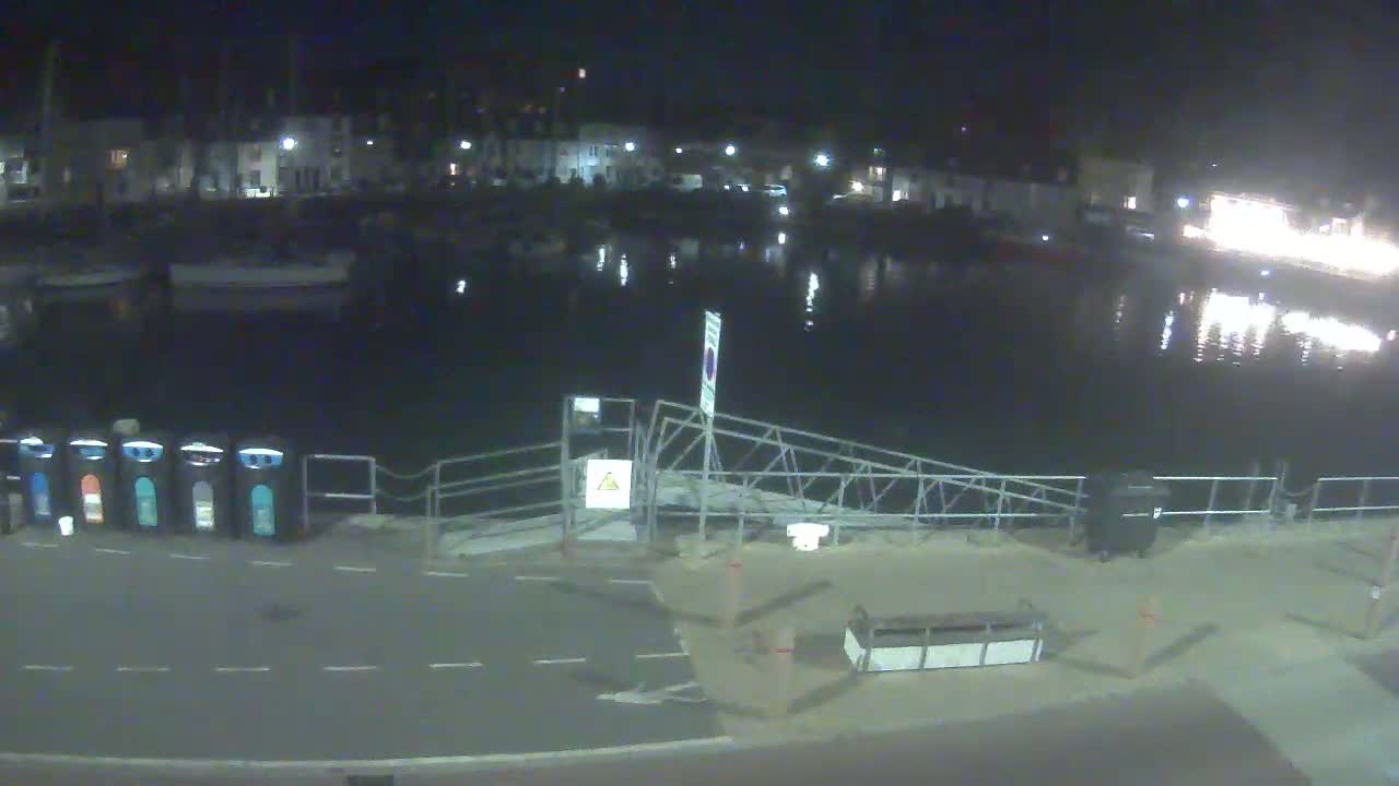 A nighttime view of a calm harbor with several boats moored, a metal gangway leading to a paved area with trash cans, and buildings illuminated in the background.