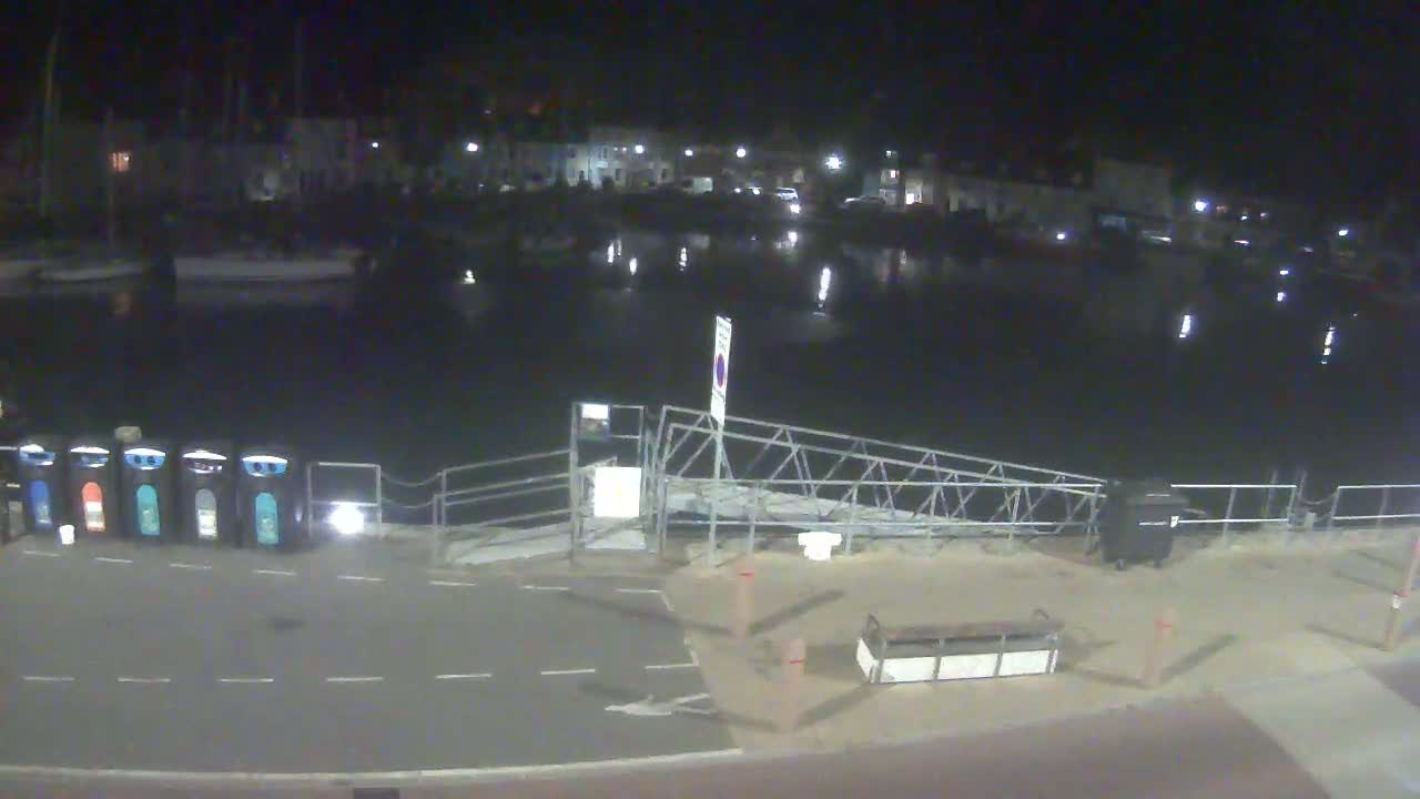 A nighttime view of a calm harbor with boats docked, a metal gangway, and several recycling bins near a paved area.