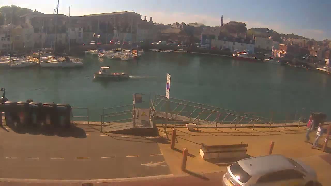 A sunny harbor scene shows several boats moored, a small boat underway, and a few people walking near a ramp beside a paved area; a silver car is partially visible in the lower right.