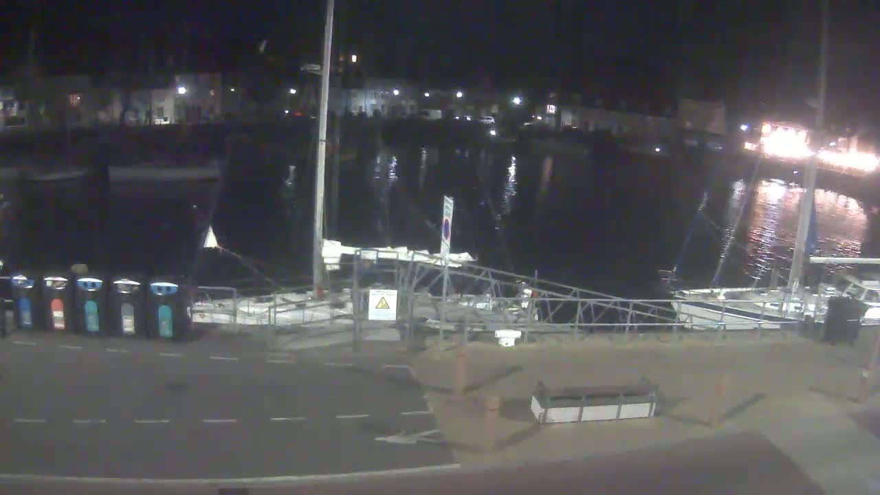 A nighttime view of a harbor with several boats docked, calm water reflecting lights from nearby buildings, and a metal gangway leading to a paved area with recycling bins.