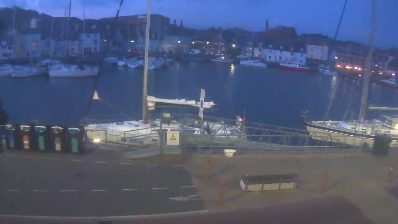 A harbor at dusk with many sailboats docked, some lights visible on buildings in the background.