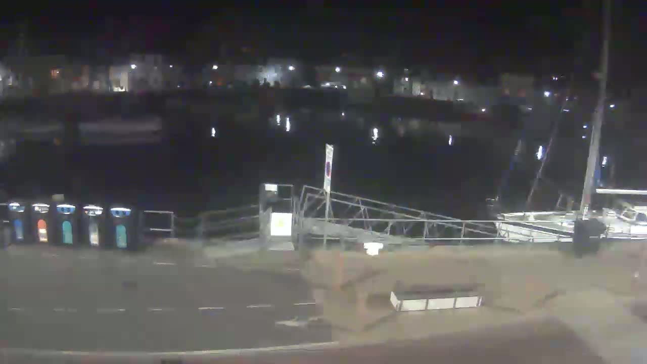A nighttime view of a calm harbor with boats moored, a metal walkway leading to a dock, and several recycling bins on the shore.