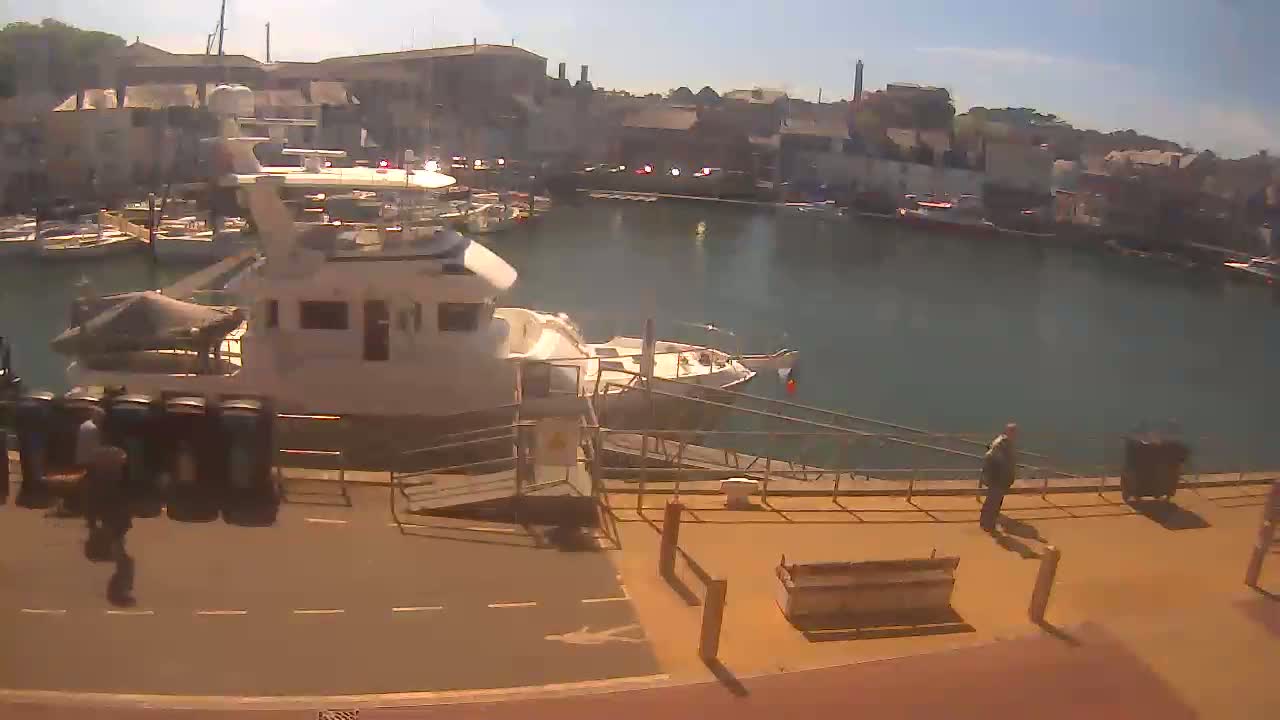 A sunny harbor scene shows a large yacht docked near a walkway with several people walking by,  smaller boats in the water, and buildings lining the harbor.