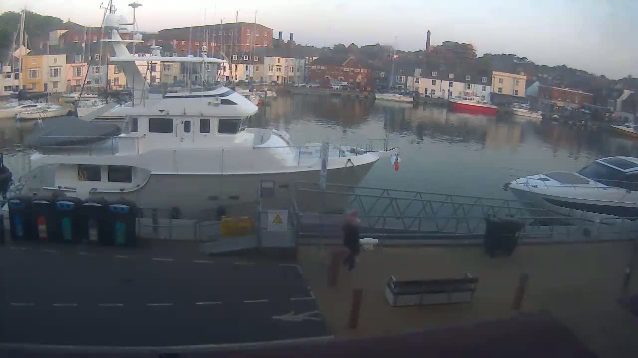 A large white motorboat is docked at a marina on a calm, overcast day, with other boats and colorful buildings visible in the background.