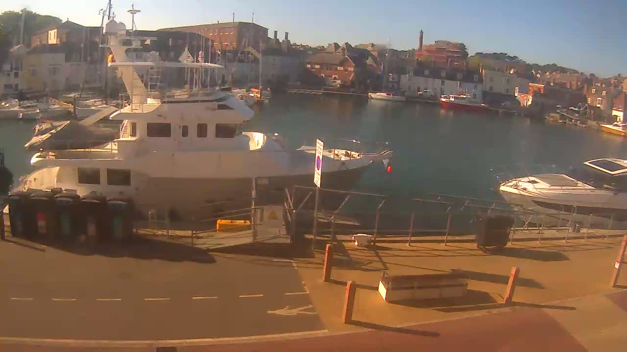 A sunny harbor scene features several boats docked alongside a paved walkway, with buildings visible in the background.