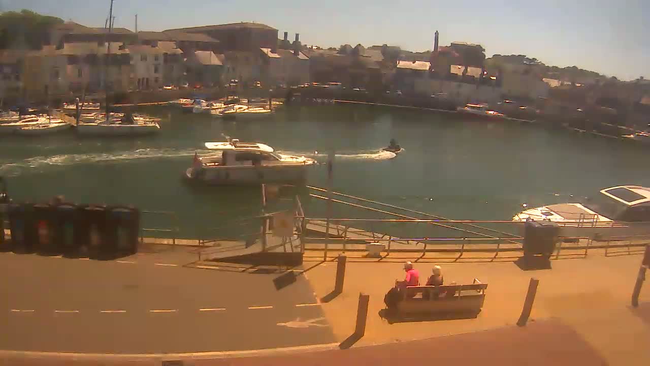 A sunny harbor scene shows several boats, a jet ski, and two people sitting on a bench near a paved walkway, with buildings lining the harbor in the background.
