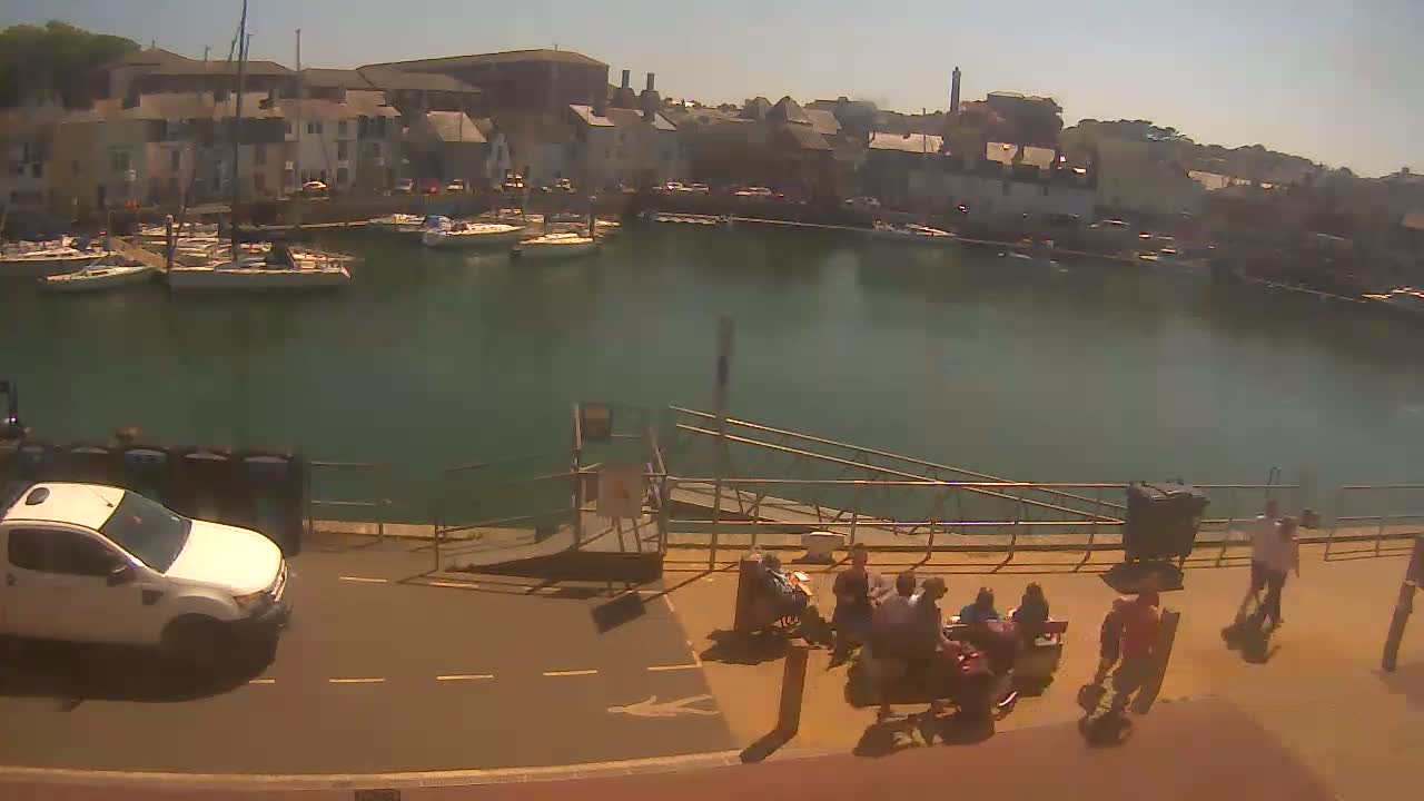 A sunny day shows a harbor filled with sailboats, a few people sitting on a dock, and a white pickup truck parked nearby.