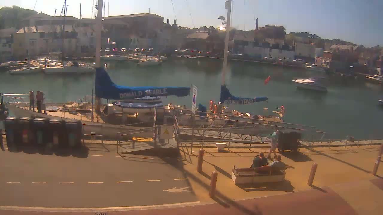 A sunny harbor scene shows numerous boats docked along buildings, with people gathered near a boat ramp on a paved area.
