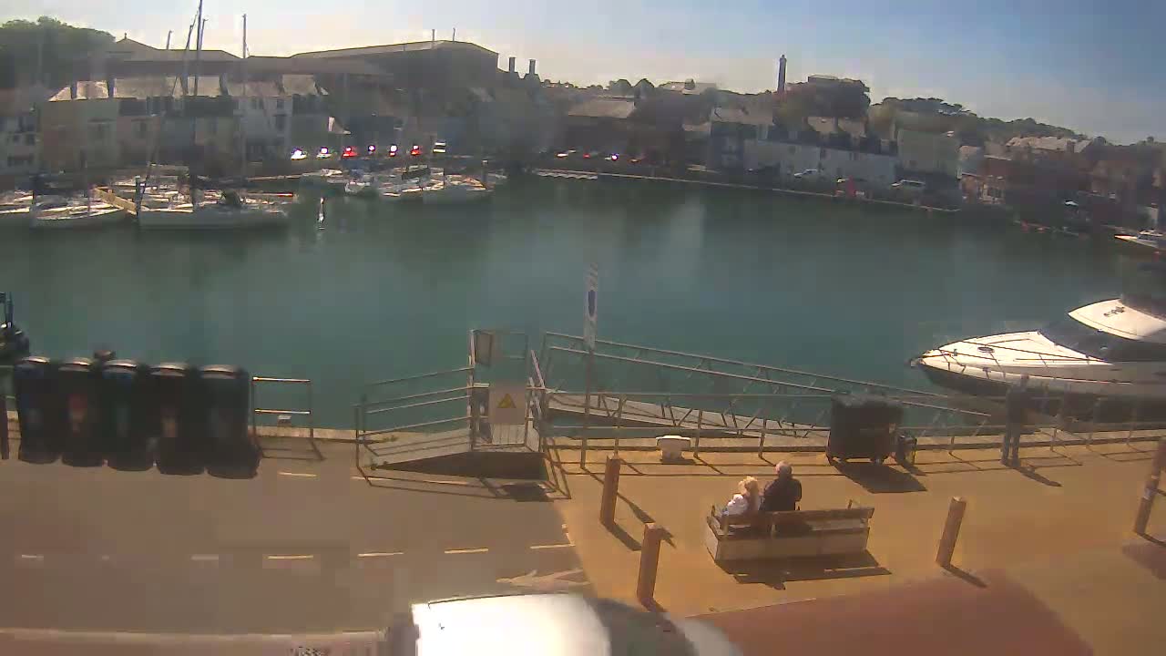 A sunny harbor scene shows a calm body of water with sailboats and a motorboat, a dock with a ramp, and a couple sitting on a bench.