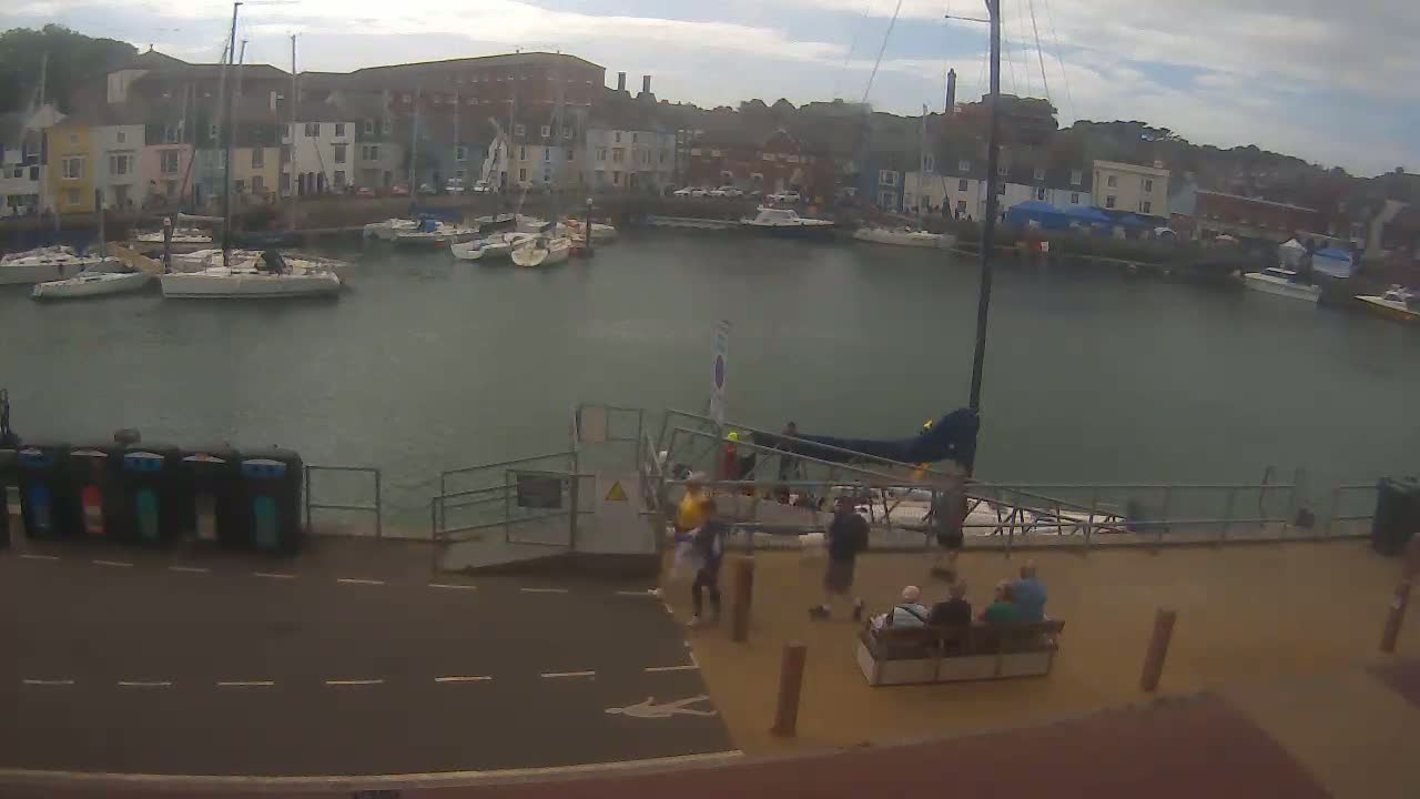 A harbor filled with sailboats is seen on an overcast day, with people gathered near a gangway leading to a sailboat.