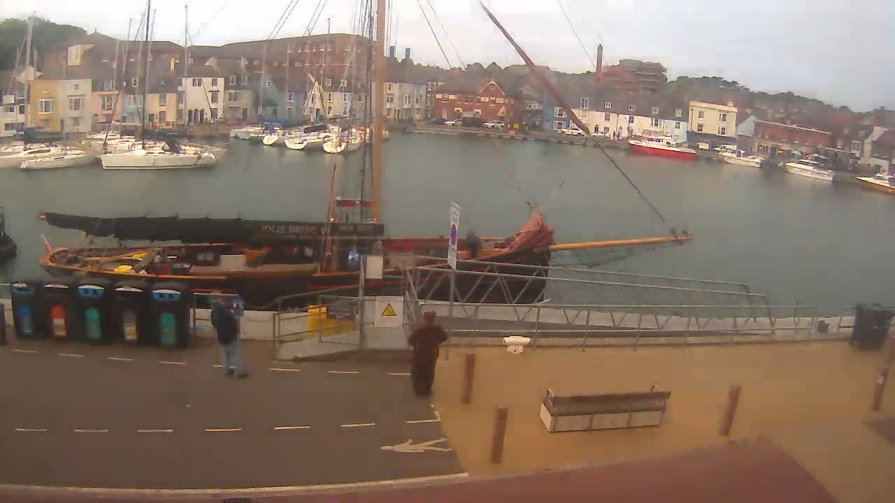 A large wooden sailing ship is docked at a harbor filled with smaller boats under an overcast sky.
