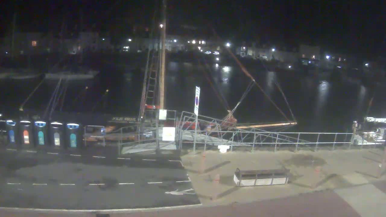 A docked sailboat is illuminated at night next to a paved area, with calm water and faintly visible lights in the background.