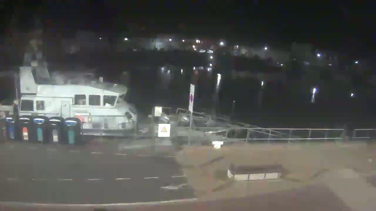 A large, light-colored boat is docked at night near a dark body of water, with a roadway and some outdoor furniture visible in the foreground.