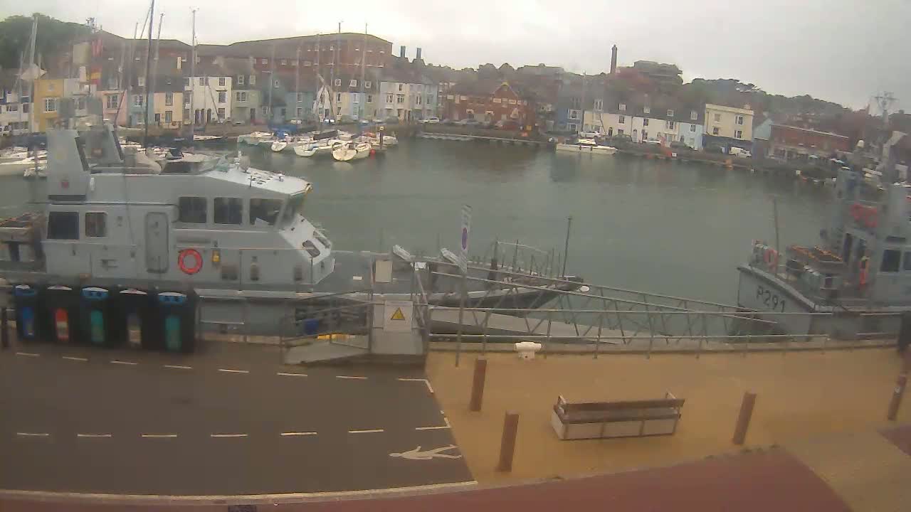 A harbor filled with docked sailboats is viewed from a quay, where two patrol boats are moored under an overcast sky.
