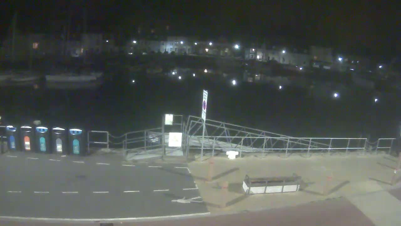A nighttime view of a calm harbor with boats moored, a small ramp leading to a paved area with several trash receptacles, and buildings illuminated in the background.