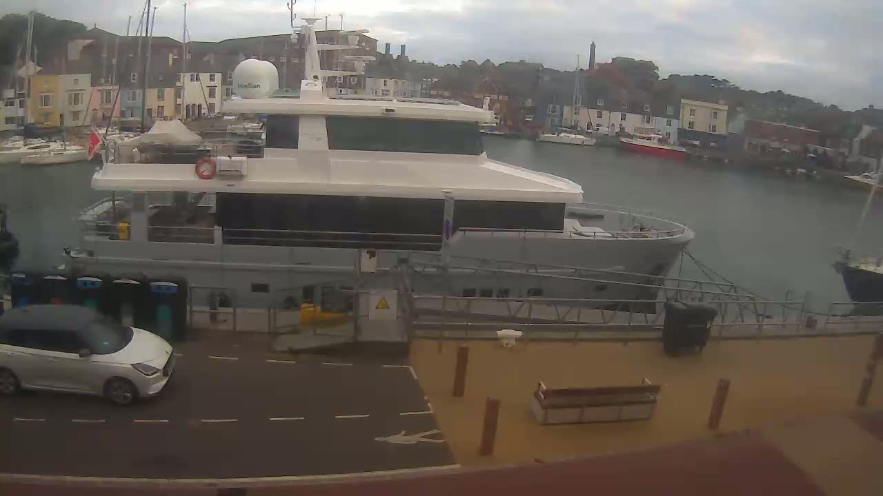A large motor yacht is docked at a marina on an overcast day, with a small white car parked nearby.
