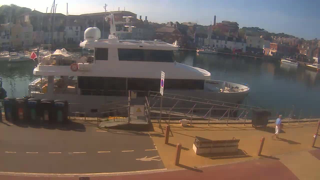 A large, light-grey motor yacht is docked at a harbor on a sunny day, with other boats and buildings visible in the background.