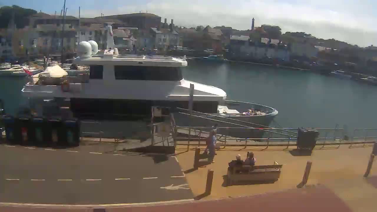 A large white motor yacht is docked at a harbor on a sunny day, with several people walking and sitting on the adjacent dock.