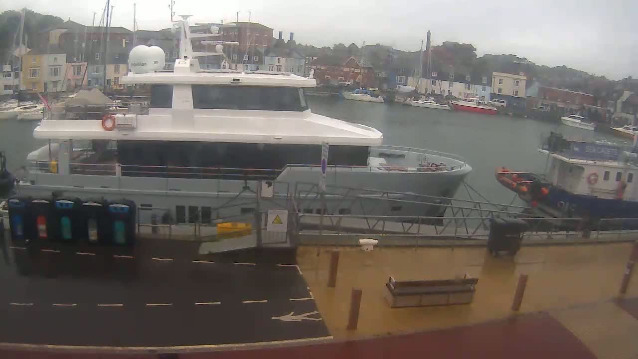 A large white and gray motor yacht is docked at a harbor on a rainy day, with other boats and buildings visible in the background.
