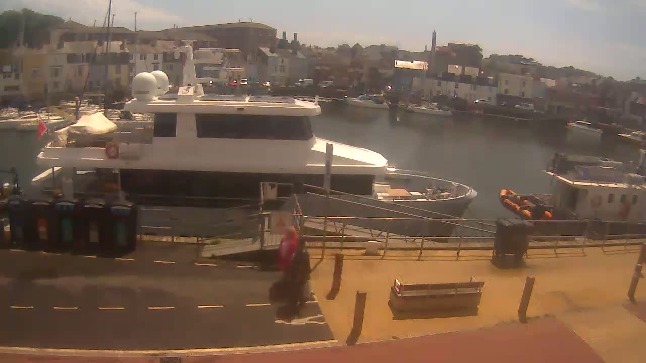 A large yacht is docked at a harbor on a sunny day, with smaller boats and buildings in the background.