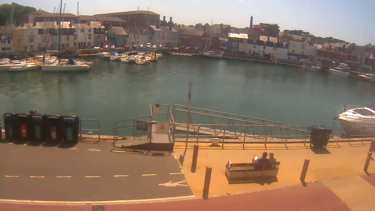A harbor filled with sailboats is viewed from above on a sunny day, with a couple sitting on a bench near a ramp leading to the water.