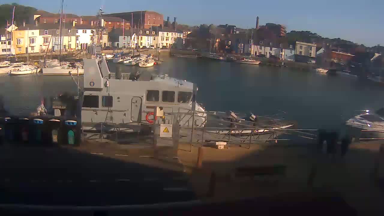 A gray patrol boat is docked in a harbor filled with sailboats and other vessels, under a sunny sky.