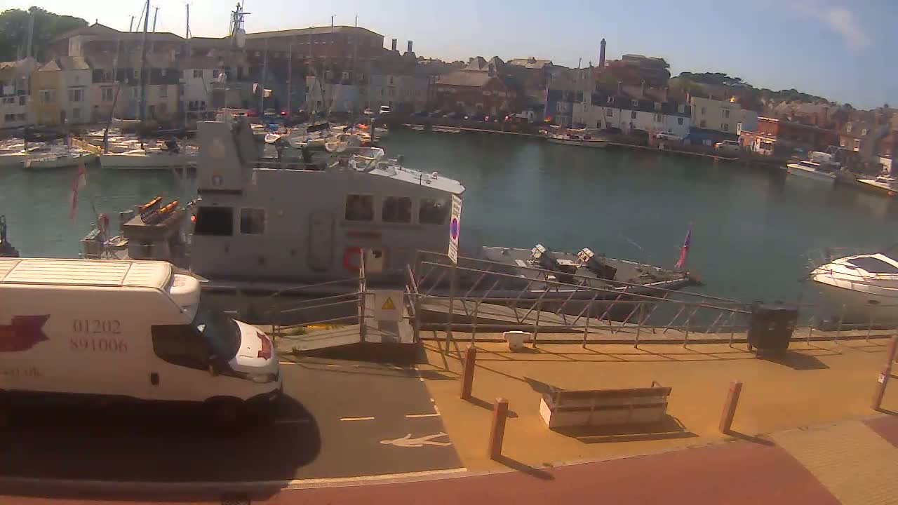 A gray patrol boat is docked in a harbor filled with other boats, under sunny skies, next to a white van and a paved walkway.