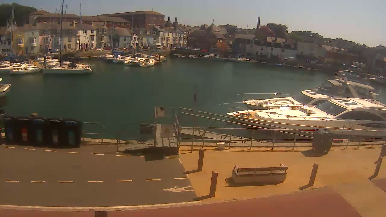 A harbor filled with sailboats and motorboats on a sunny day, viewed from a waterfront walkway.
