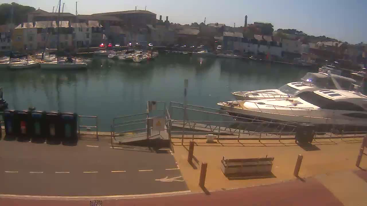 A marina filled with sailboats and motorboats under sunny skies, with two large motor yachts docked at a ramp near a paved walkway.