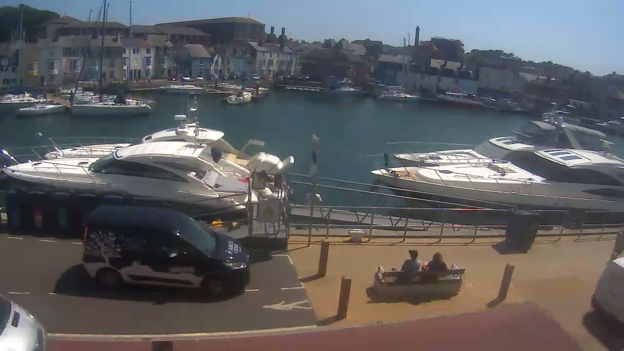 A sunny day at a marina shows several docked boats, a van parked nearby, and two people sitting on a bench.