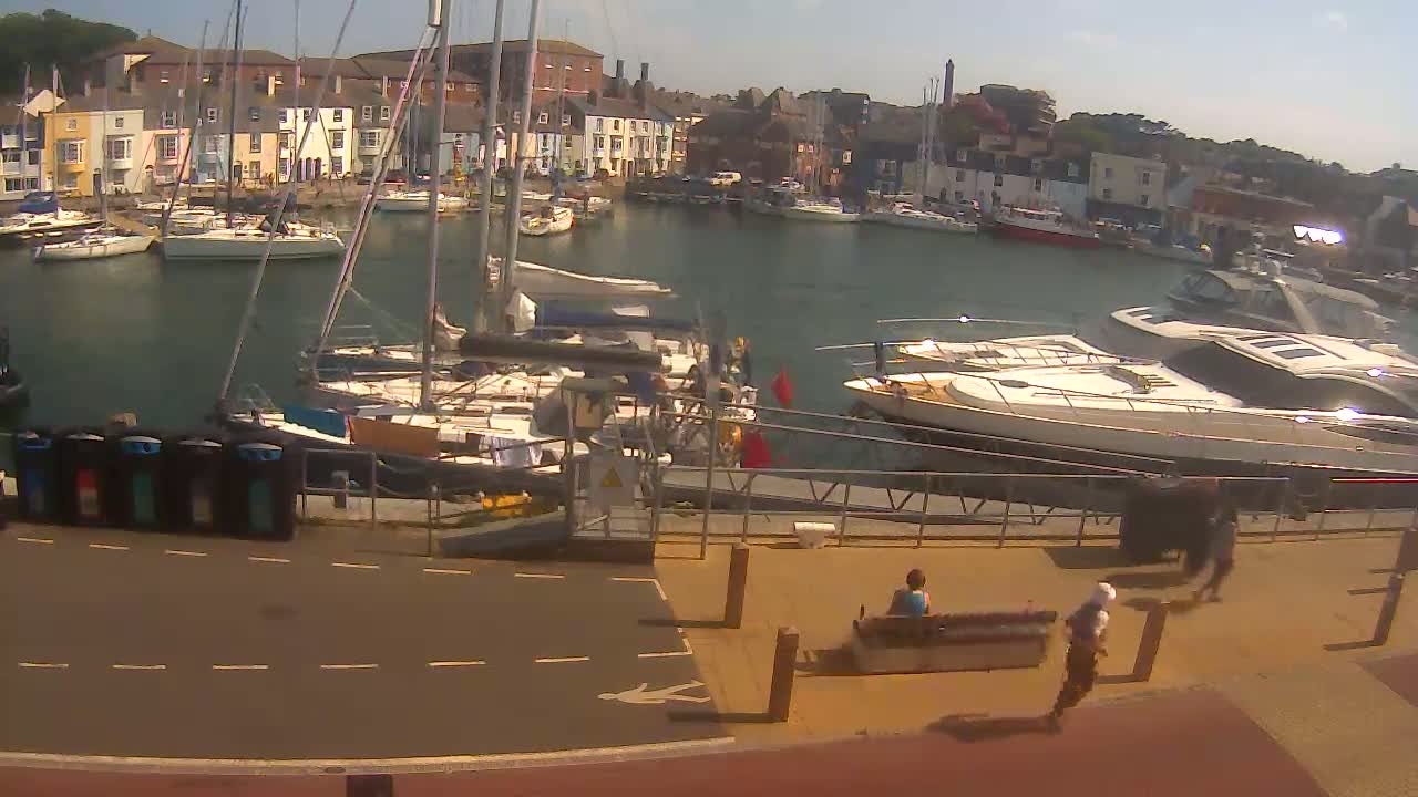 A harbor filled with sailboats and motorboats on a sunny day, with a paved walkway and people walking nearby.