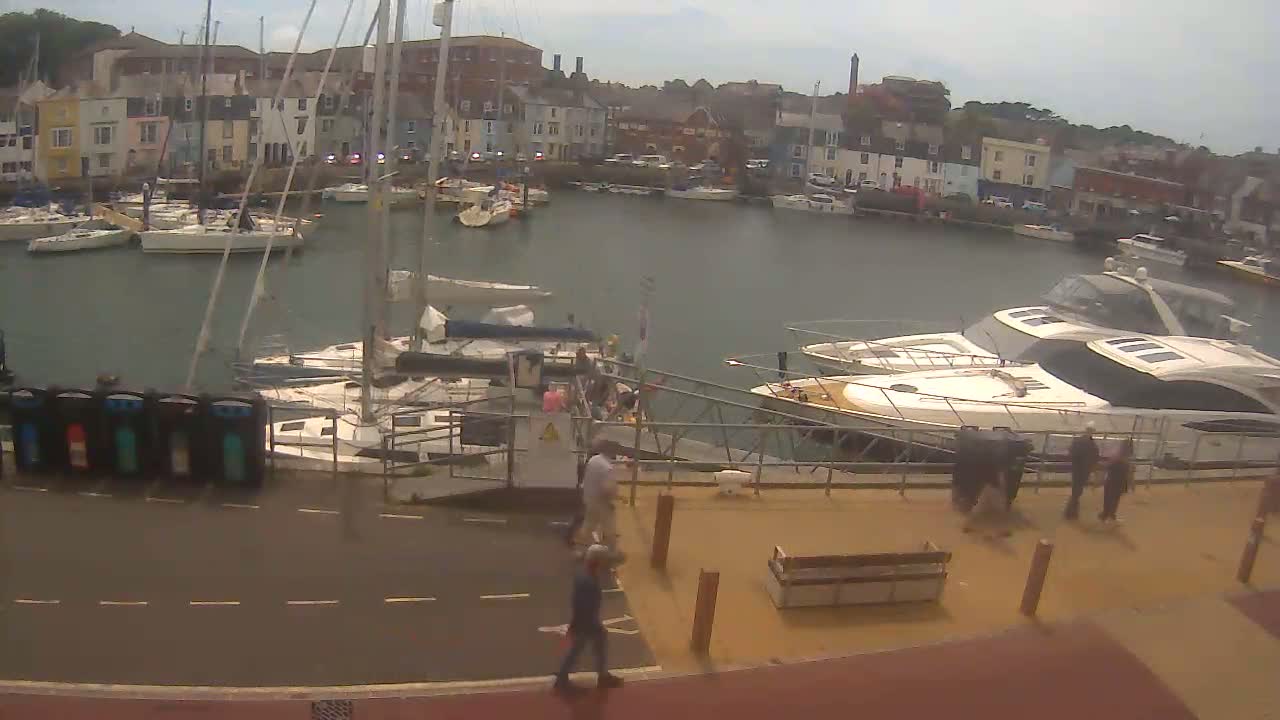 A harbor filled with sailboats and motorboats on an overcast day, with several people walking along a nearby paved area.