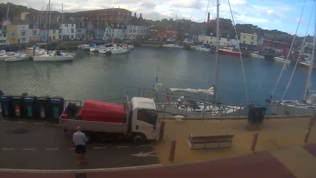 A harbor filled with sailboats is viewed from above on an overcast day, with a small truck parked on a nearby road.