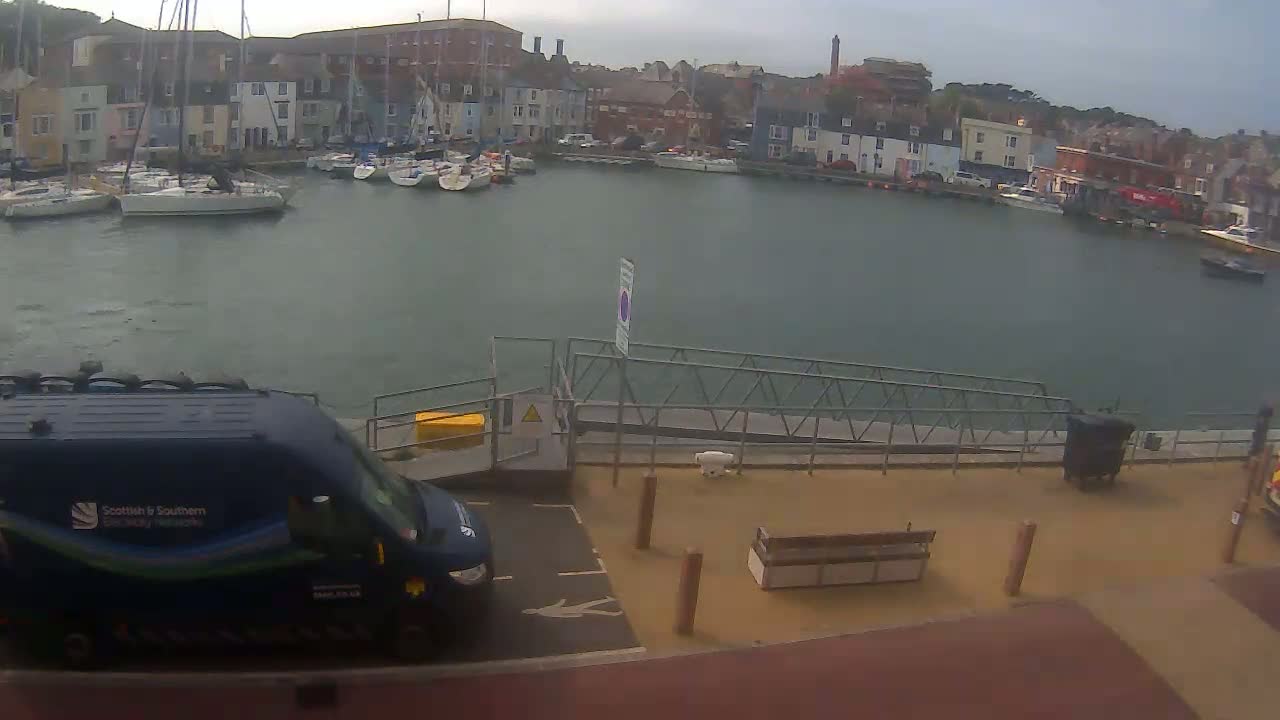A harbor filled with sailboats is viewed from a quay where a blue van is parked near a bench under an overcast sky.