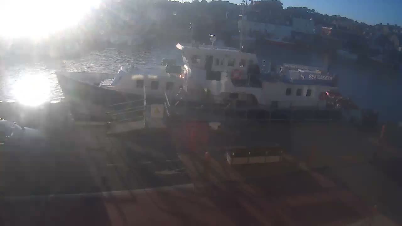 A bright, sunny day illuminates a harbor where several boats are docked along the shore, with strong sun glare on the water and buildings lining a distant hillside.