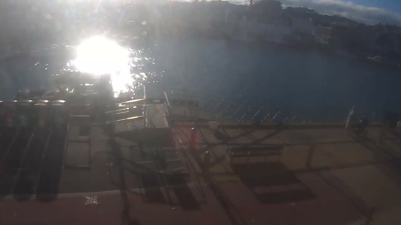 A bright, sunny day illuminates a harbor where several boats are docked along the shore, with strong sun glare on the water and buildings lining a distant hillside.
