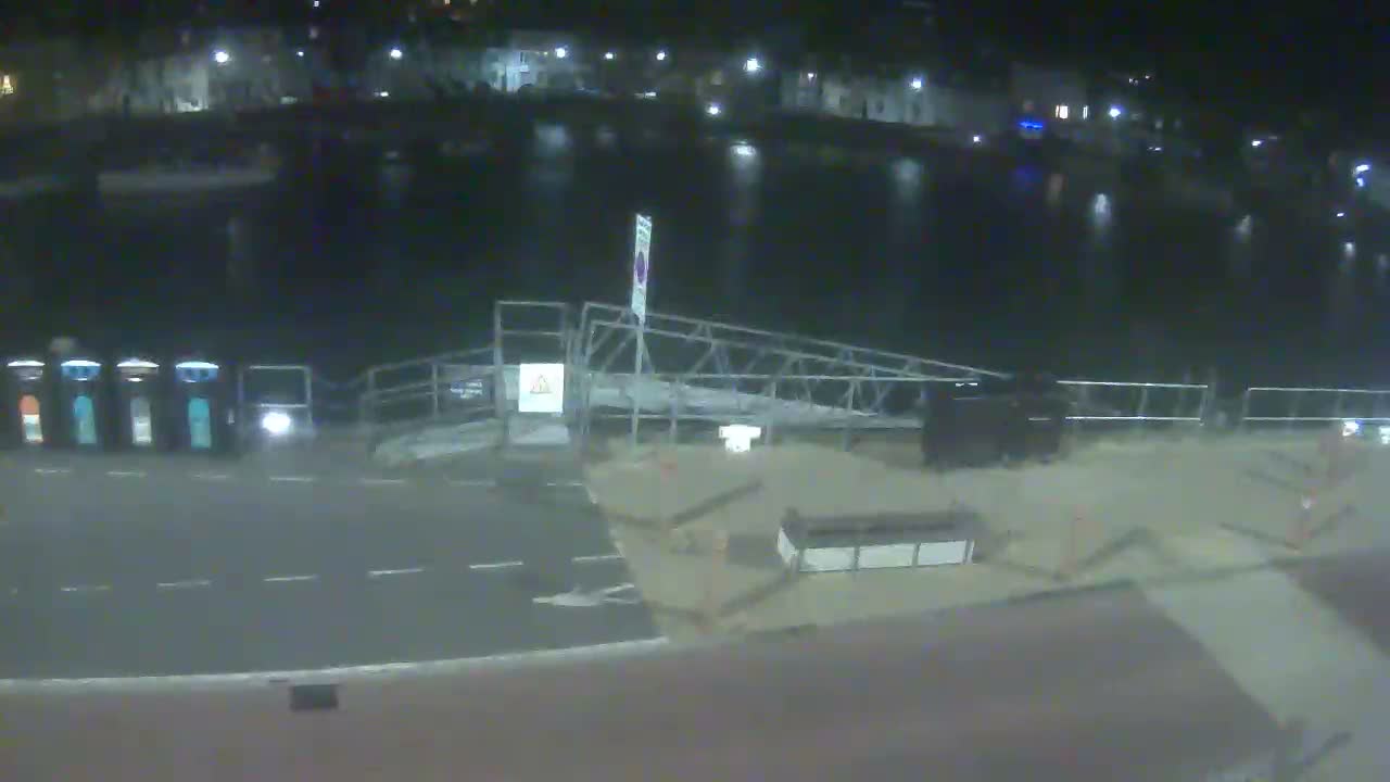An overcast sky hangs over a calm harbor filled with numerous moored boats and bordered by town buildings, as seen from an elevated perspective overlooking a quay with portable toilets and a gangway.