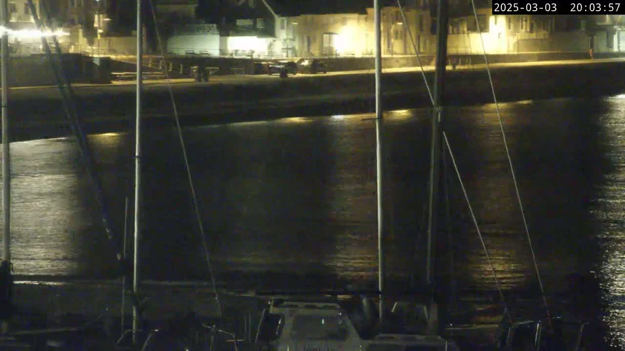 At night, a calm body of water reflects lights from a nearby shoreline and several boats.