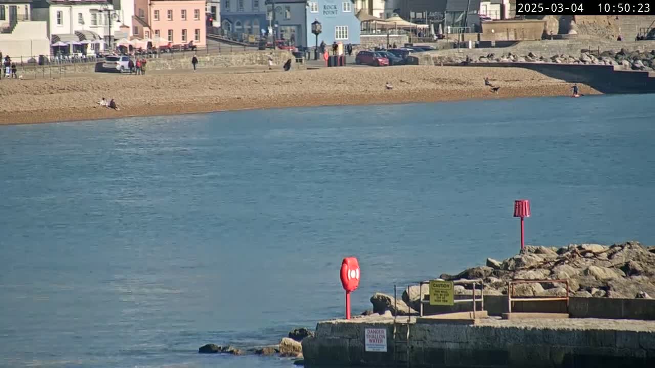 A sunny day shows a calm body of water next to a sandy beach with people and buildings in the background.