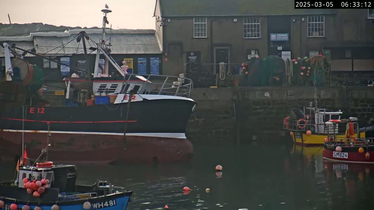 Several fishing boats are moored in a harbor next to a stone building on a calm, overcast day.