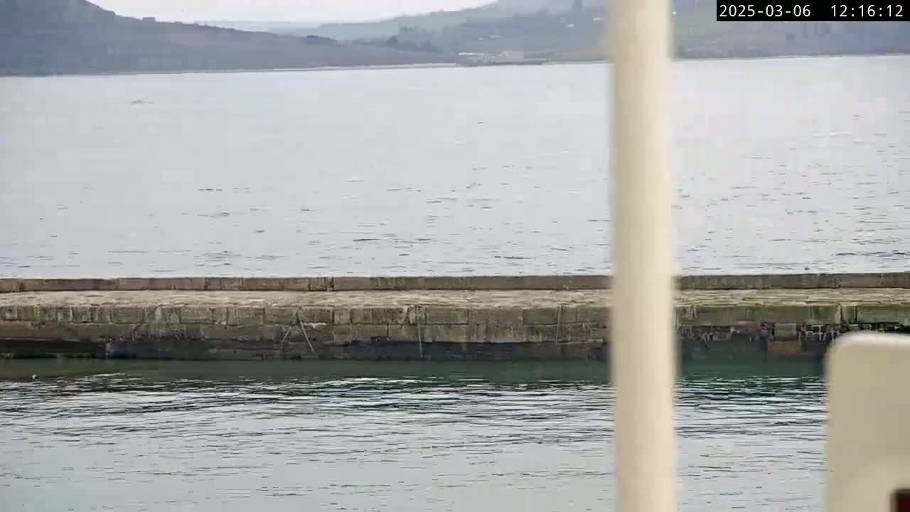 A low stone wall separates calm, gray water from a distant, hazy shoreline.